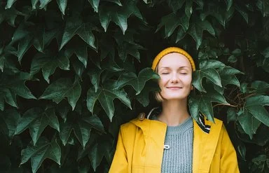 Ein Frau steht mit geschlossenen Augen in einem gelben Regenmantel vor einen Blätterwand.
