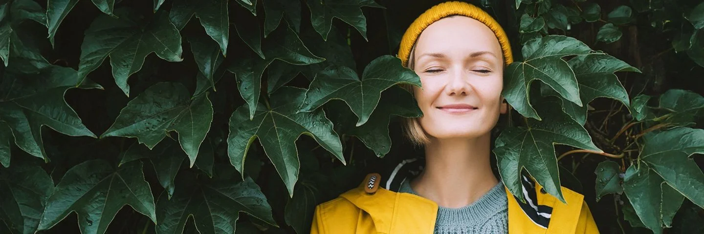 Ein Frau steht mit geschlossenen Augen in einem gelben Regenmantel vor einen Blätterwand.