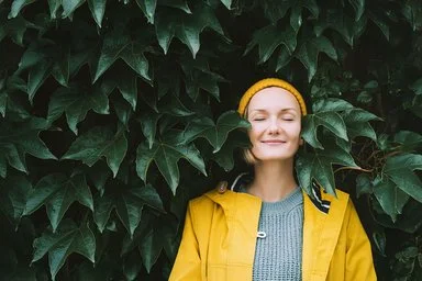 Ein Frau steht mit geschlossenen Augen in einem gelben Regenmantel vor einen Blätterwand.