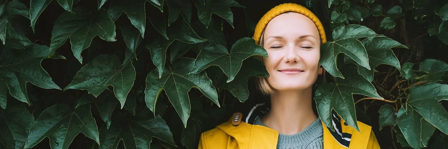 Ein Frau steht mit geschlossenen Augen in einem gelben Regenmantel vor einen Blätterwand.