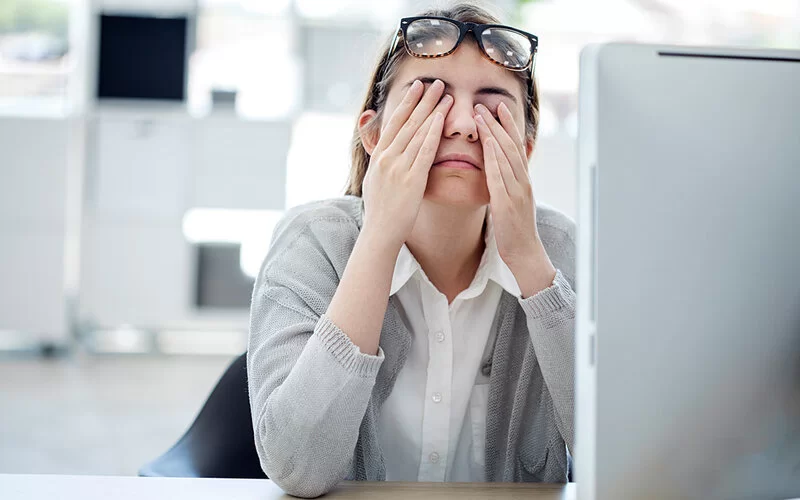 Eine junge Frau sitzt im Büro vor ihrem Rechner und berührt sanft ihre geschlossenen Augen.