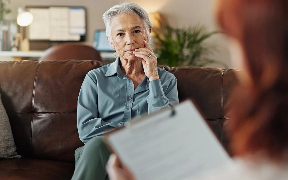 Eine Frau sitzt auf dem Sofa im Behandlungszimmer einer Therapeutin. Sie schaut die Therapeutin konzentriert an, die nur von hinten zu sehen ist und ein Klemmbrett in der Hand hält.