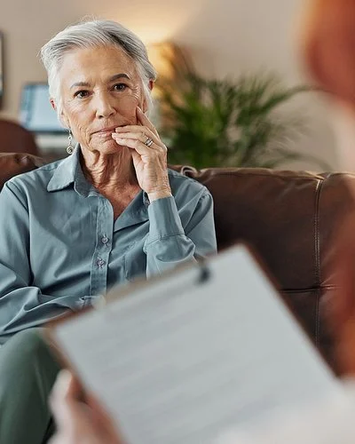 Eine Frau sitzt auf dem Sofa im Behandlungszimmer einer Therapeutin. Sie schaut die Therapeutin konzentriert an, die nur von hinten zu sehen ist und ein Klemmbrett in der Hand hält.