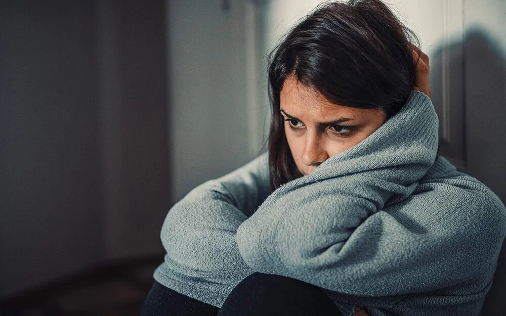 Nahaufnahme einer unglücklich wirkenden Frau. Sie sitzt an die Wand gelehnt auf dem Fußboden, ihre Ellenbogen ruhen auf den angezogenen Knien, die linke Wange ist an ihren linken Unterarm geschmiegt.