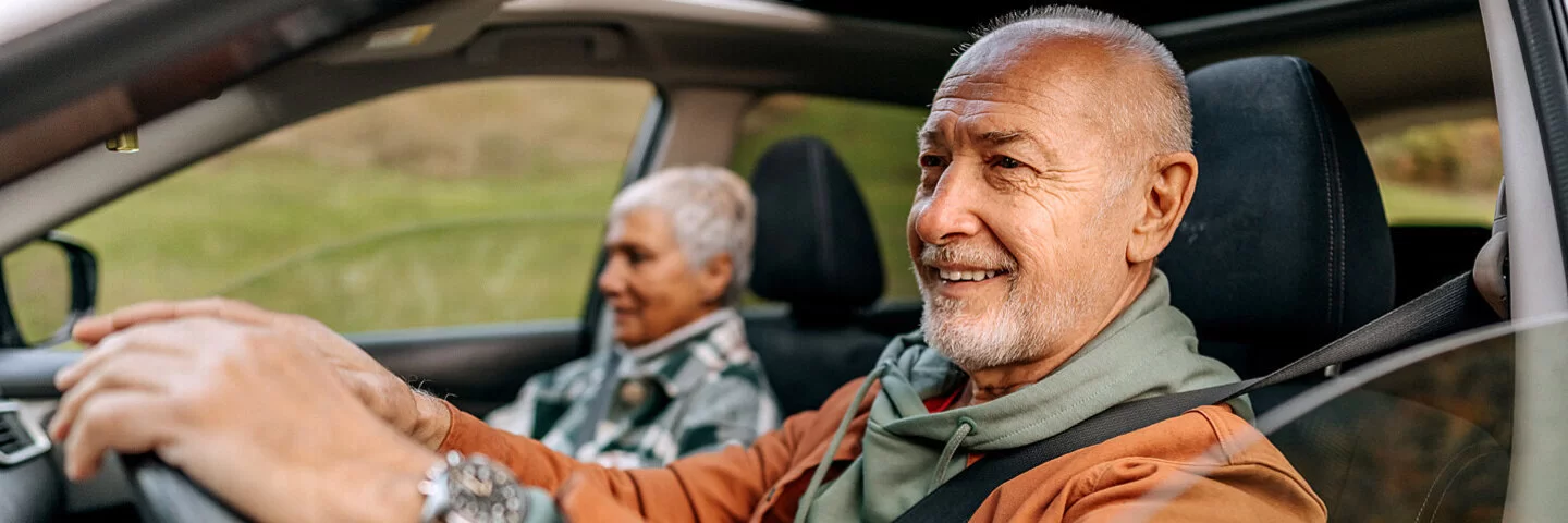 Ein Seniorenpaar sitzt angeschnallt im Auto, er hinterm Lenkrad und sie auf dem Beifahrersitz.