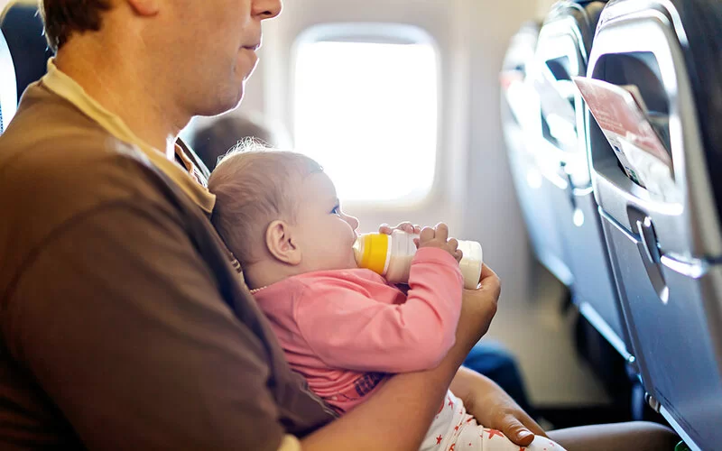 Ein Baby sitzt im Flugzeug auf dem Schoß des Vaters und nuckelt an einer Trinkflasche.