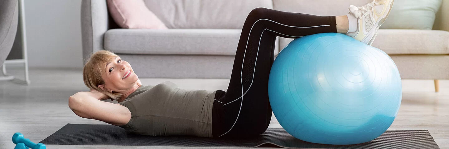 Eine ältere Frau liegt rücklings auf einer Yoga Matte und hat ihre Unterschenkel auf einem Gymnastikball vor sich abgelegt.