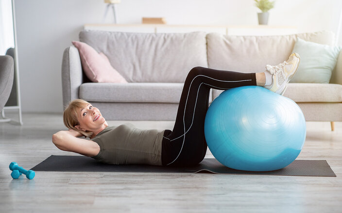 Eine ältere Frau liegt rücklings auf einer Yoga Matte und hat ihre Unterschenkel auf einem Gymnastikball vor sich abgelegt.