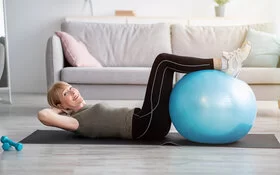 Eine ältere Frau liegt rücklings auf einer Yoga Matte und hat ihre Unterschenkel auf einem Gymnastikball vor sich abgelegt.