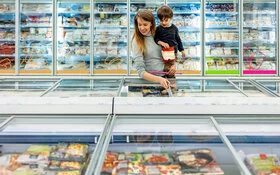 Eine Frau und ihr Sohn auf dem Arm sind in einem Supermarkt und suchen in den Tiefkühltruhen nach gesunden und klimaschonenden Lebensmitteln.