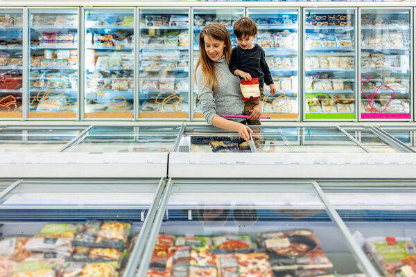 Eine Frau und ihr Sohn auf dem Arm sind in einem Supermarkt und suchen in den Tiefkühltruhen nach gesunden und klimaschonenden Lebensmitteln.