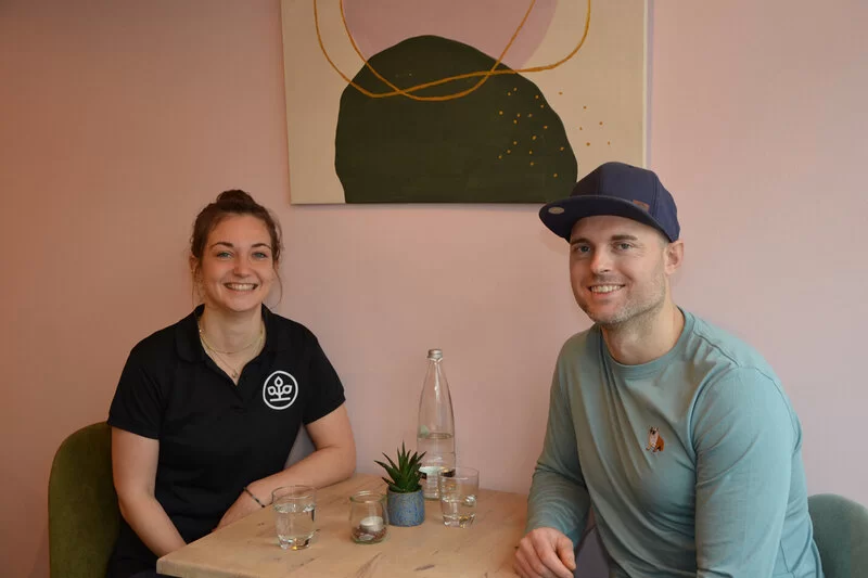 Die Moderatoren der AOK-Kochshow Mannheim sitzen an einem Tisch und lächeln in die Kamera.