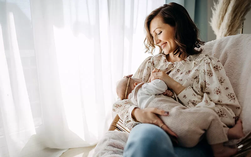 Eine junge Mutter sitzt gemütlich mir ihrem Baby in einem Korbstuhl und stillt es. Im Hintergrund scheint die Sonne durch transparente Vorhänge.