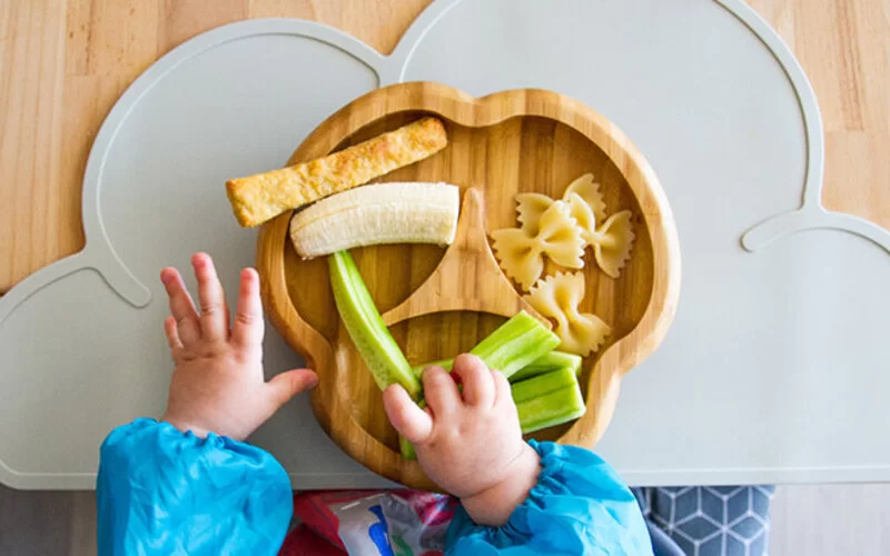 Baby sitzt im Hochstuhl und greift zu einem Stück Gurke, da es bereits an feste Nahrung gewöhnt ist