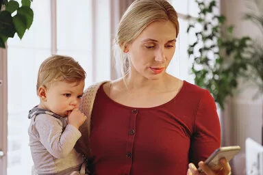 Eine etwa 30-jährige Frau mit blonden zusammengebundenen Haaren, weinrotem T-Shirt und weißem Cardigan steht mit ihrem Smartphone in der Hand im Wohnzimmer und hält ihr Baby im anderen Arm.