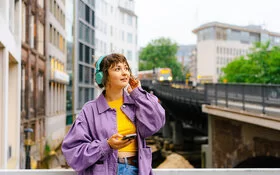 Eine Frau mit türkisfarbenen Kopfhörern steht auf einer Brücke, hinter ihr fährt ein Zug entlang.