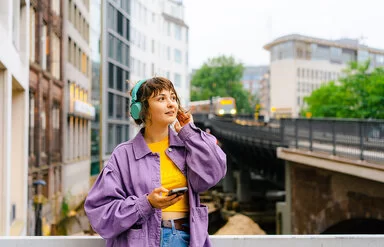 Eine Frau mit türkisfarbenen Kopfhörern steht auf einer Brücke, hinter ihr fährt ein Zug entlang.