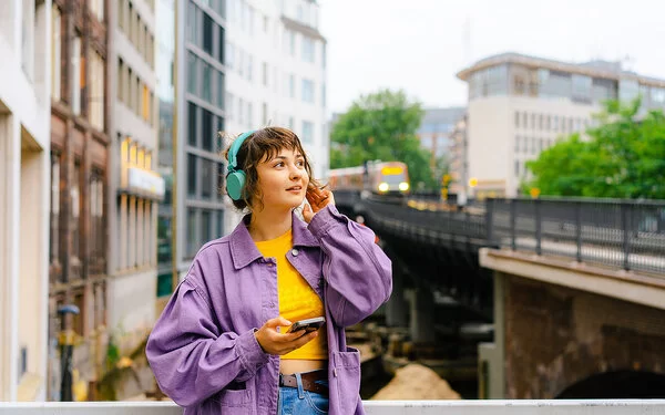 Eine Frau mit türkisfarbenen Kopfhörern steht auf einer Brücke, hinter ihr fährt ein Zug entlang.