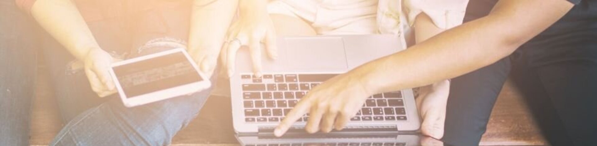 Vier Menschen sitzen nebeneinander auf einer Bank. Zwei von ihnen haben einen Laptop auf dem Schoß, eine weitere hält ein Tablet in den Händen. Die vierte Person zeigt auf einen der Bildschirme. Der Bildausschnitt zeigt lediglich die Schöße der Personen.