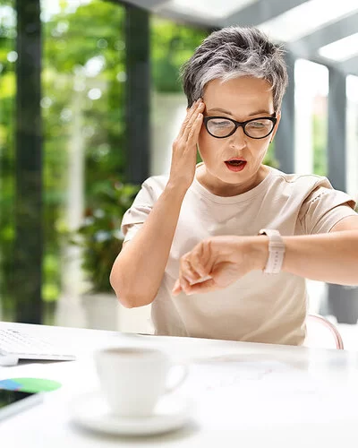 Eine Frau mit leicht grauen Haaren und Brille sitzt an einem Schreibtisch, vor sich Monitor, Tastatur und einige Unterlagen. Sie blickt auf ihre Armbanduhr, fasst sich an die Stirn und schaut erschrocken.