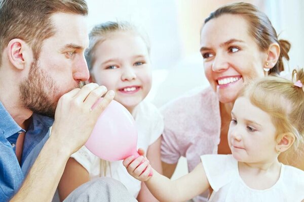 Eine Familie mit zwei kleinen Kindern schaut dem Vater dabei zu, wie er einen Luftballon aufbläst.