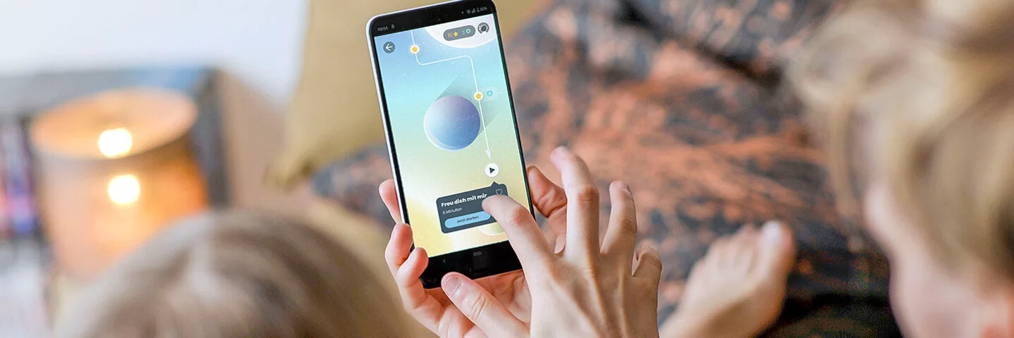 Zwei nicht erkennbare Kinder sitzen mit dem Rücken zur Kamera auf einem Bett. Eines der Kinder hält ein Smartphone mit der Aumio-App in der Hand, das andere Kind tippt aufs Display.