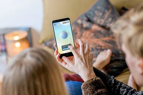 Zwei nicht erkennbare Kinder sitzen mit dem Rücken zur Kamera auf einem Bett. Eines der Kinder hält ein Smartphone mit der Aumio-App in der Hand, das andere Kind tippt aufs Display.