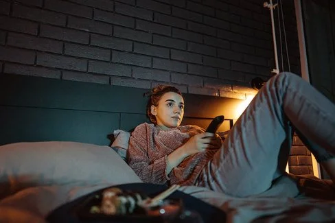 Eine junge Frau liegt mit einer Fernbedienung in der Hand auf einem Bett und schaut auf einen nicht sichtbaren Fernseher.