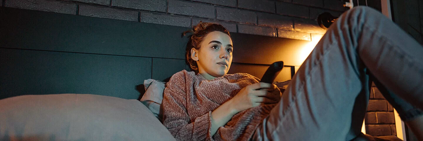 Eine junge Frau liegt mit einer Fernbedienung in der Hand auf einem Bett und schaut auf einen nicht sichtbaren Fernseher.