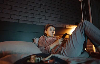 Eine junge Frau liegt mit einer Fernbedienung in der Hand auf einem Bett und schaut auf einen nicht sichtbaren Fernseher.