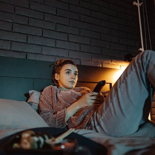 Eine junge Frau liegt mit einer Fernbedienung in der Hand auf einem Bett und schaut auf einen nicht sichtbaren Fernseher.