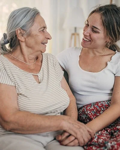 Eine ältere Frau und ihre Tochter sitzen zusammen auf einem Sofa und schauen sich freundlich an.