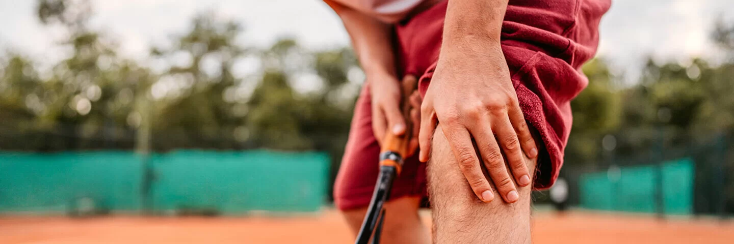 Ein männlicher Tennisspieler fasst sich auf dem Tennisplatz ans Knie, nachdem ein Meniskus gerissen ist.