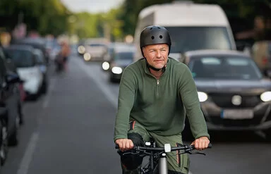 Ein Mann mit grüner Arbeitshose und grünem Pullover fährt mit dem Fahrrad auf der Fahrradspur der Straße. Neben ihm sind viele Autos zu sehen. Er trägt einen schwarzen Fahrradhelm und fährt konzentriert auf die Kamera zu.