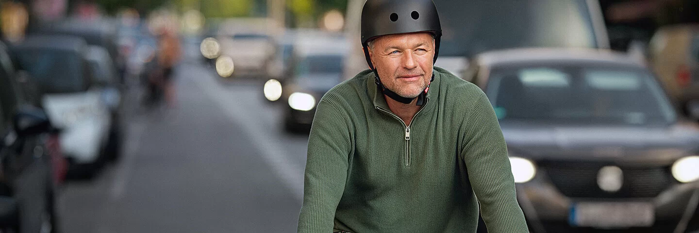 Ein Mann mit grüner Arbeitshose und grünem Pullover fährt mit dem Fahrrad auf der Fahrradspur der Straße. Neben ihm sind viele Autos zu sehen. Er trägt einen schwarzen Fahrradhelm und fährt konzentriert auf die Kamera zu.