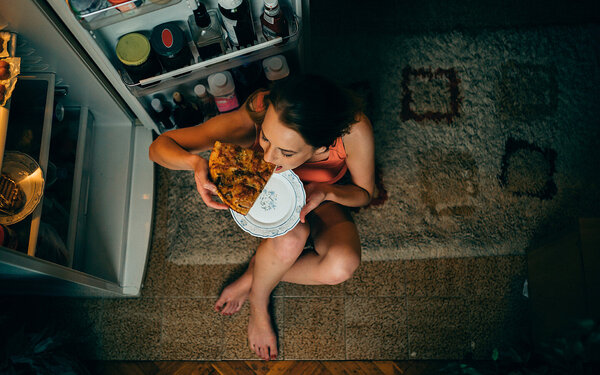 Eine Frau sitzt vor dem Kühlschrank und verzehrt einen Mitternachtssnack.