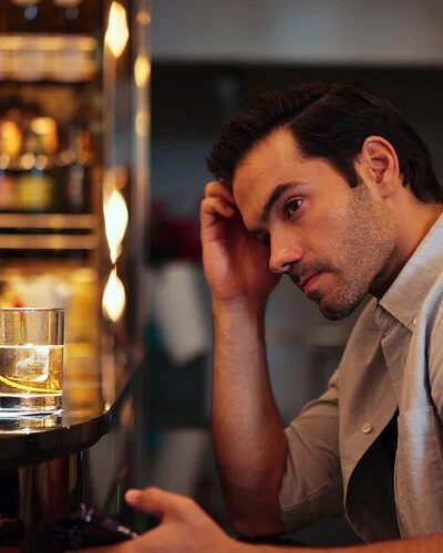 Ein dunkelhaariger Mann um die dreißig im hellen Businesshemd sitzt am Tresen einer Bar, vor sich ein Glas Whisky. Er blickt grübelnd ins Leere.