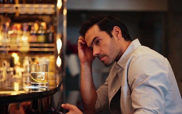 Ein dunkelhaariger Mann um die dreißig im hellen Businesshemd sitzt am Tresen einer Bar, vor sich ein Glas Whisky. Er blickt grübelnd ins Leere.