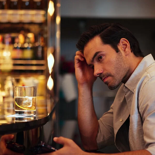 Ein dunkelhaariger Mann um die dreißig im hellen Businesshemd sitzt am Tresen einer Bar, vor sich ein Glas Whisky. Er blickt grübelnd ins Leere.
