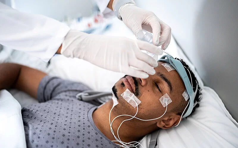 Ein junger Mann liegt auf einem Bett in einer medizinischen Einrichtung. Am Kopf sind Elektroden befestigt und er trägt ein Stirnband. Eine Person, von der nur die Unterarme und die Hände in Latexhandschuhen im Bildausschnitt zu sehen sind, bringt eine Beatmungsmaske an Stirnband und Nase des Mannes an.