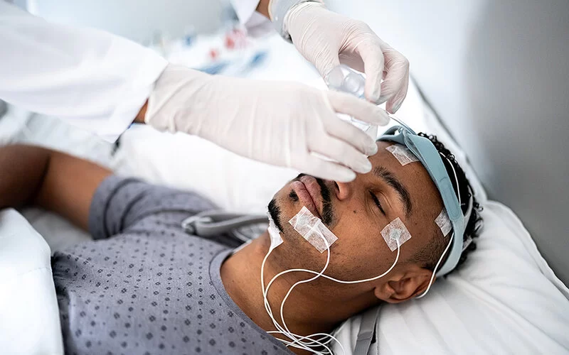 Ein junger Mann liegt auf einem Bett in einer medizinischen Einrichtung. Am Kopf sind Elektroden befestigt und er trägt ein Stirnband. Eine Person, von der nur die Unterarme und die Hände in Latexhandschuhen im Bildausschnitt zu sehen sind, bringt eine Beatmungsmaske an Stirnband und Nase des Mannes an.