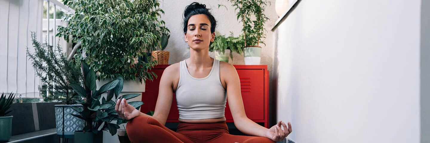 Eine junge Frau sitzt im Lotus-Sitz auf einem Balkon auf einer Yoga-Matte und hält die Augen geschlossen.