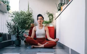 Eine junge Frau sitzt im Lotus-Sitz auf einem Balkon auf einer Yoga-Matte und hält die Augen geschlossen.