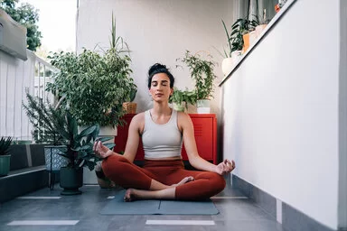 Eine junge Frau sitzt im Lotus-Sitz auf einem Balkon auf einer Yoga-Matte und hält die Augen geschlossen.