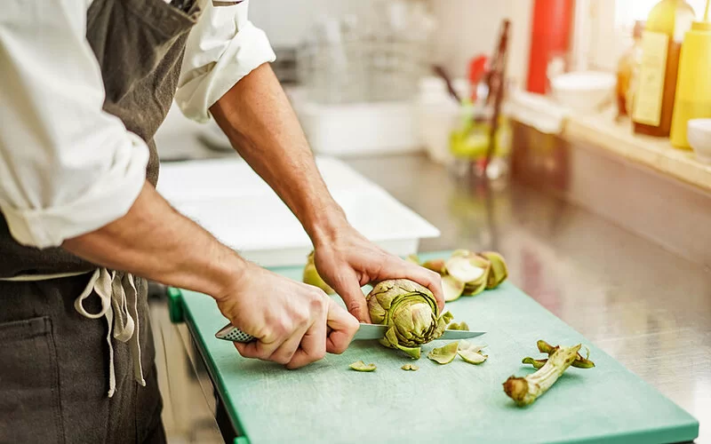 Koch schneidet Artischocke auf einem grünen Brett. Er befindet sich in einer Restaurant-Küche.