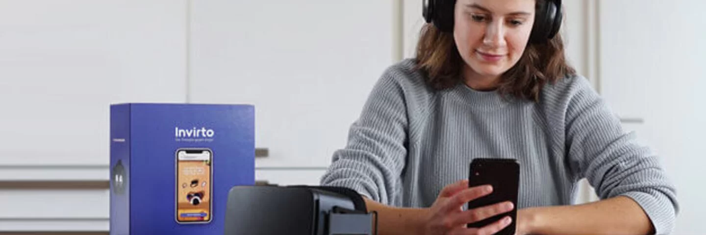 Eine junge Frau sitzt an einem Tisch, trägt Kopfhörer und schaut auf ein Handy, das sie in ihrer rechten Hand hält. Neben ihr auf dem Tisch liegen eine schwarze Virtual-Reality-Brille und eine violette Box, auf der die "Invirto"-App auf einem Smartphone abgebildet ist.