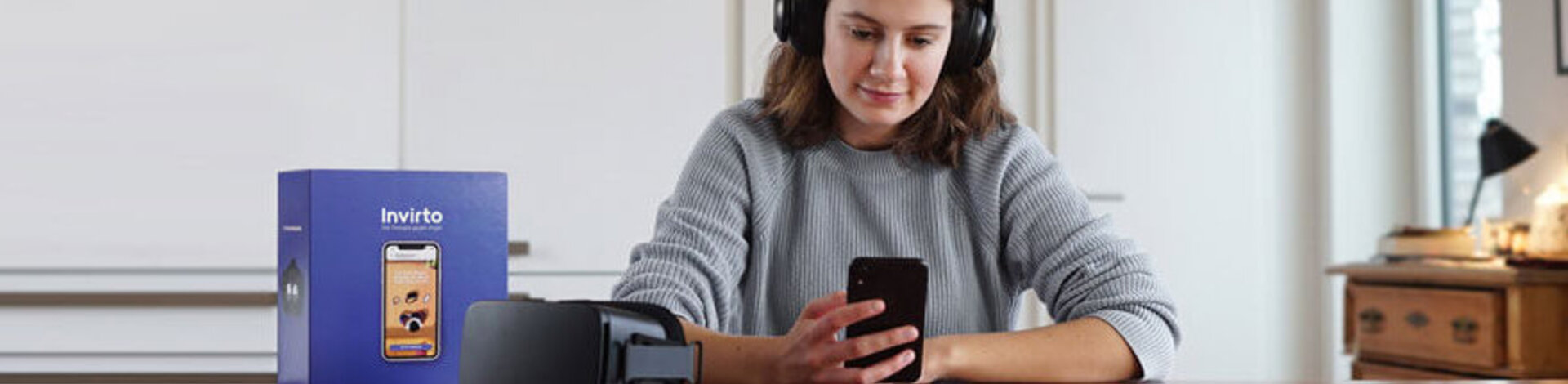 Eine junge Frau sitzt an einem Tisch, trägt Kopfhörer und schaut auf ein Handy, das sie in ihrer rechten Hand hält. Neben ihr auf dem Tisch liegen eine schwarze Virtual-Reality-Brille und eine violette Box, auf der die "Invirto"-App auf einem Smartphone abgebildet ist.
