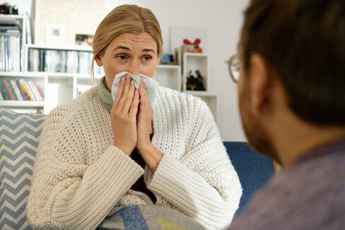 Eine Frau ist zu Hause und putzt ihre Nase. Falls sie aufgrund eines grippalen Infekts eine Krankschreibung erhalten hat, sollte sie das Dokument schnell an die AOK schicken. Zukünftig geschieht das mit der eAU elektronisch direkt vom Arzt aus.