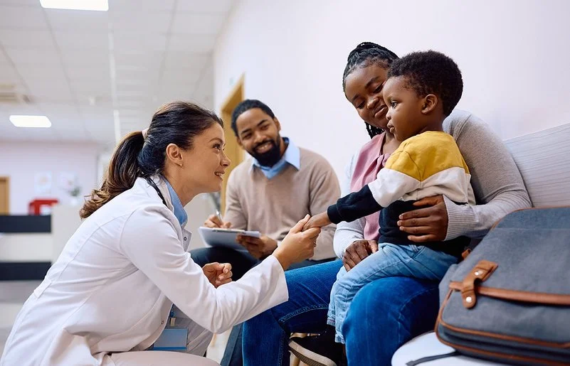 Eine junge Ärztin interagiert mit einem Kind und seiner Mutter im Warteraum einer Klinik.
