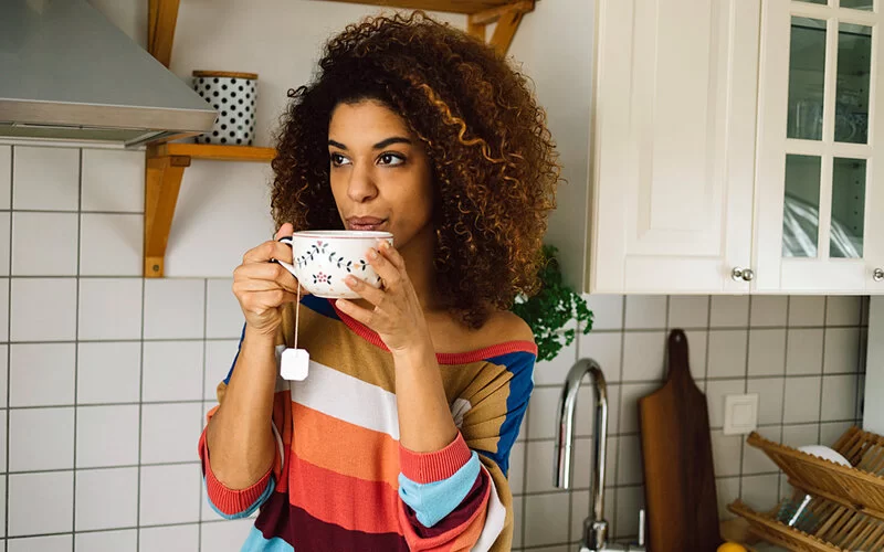 Junge Frau steht in der Küche und pustet auf eine Tasse mit heißem Tee.